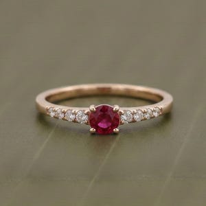 May include: A rose gold ring featuring a central round-cut red ruby gemstone held by four prongs. The band is accented with three small, clear, round-cut white diamonds on each side of the center stone, creating a symmetrical design.