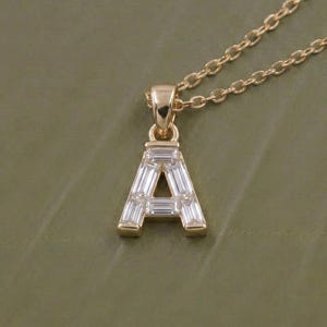 以下が含まれることがあります： ゴールドカラーのネックレス。ペンダントは文字「A」の形をしており、長方形の透明な宝石がちりばめられています。チェーンもゴールドカラーで繊細です。背景は落ち着いた緑色です。