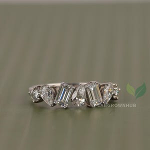 May include: A silver ring featuring a series of clear, sparkling gemstones in various shapes, including pear and emerald cuts. The ring is set against a muted green background, showcasing the brilliance of the stones.