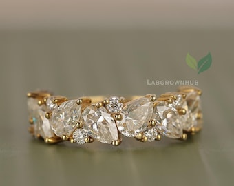 May include: A gold ring featuring a series of pear-shaped and round clear gemstones. The ring has a delicate design with the gemstones set closely together, creating a continuous pattern. The ring is set against a soft, muted background.