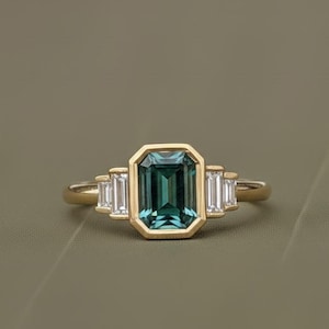 Puede incluir: Un anillo de oro con una piedra preciosa de color verde azulado de talla esmeralda en el centro, flanqueada por dos piedras preciosas rectangulares y transparentes más pequeñas a cada lado. El anillo tiene un diseño sencillo y elegante.