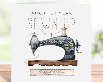 May include: A square greeting card featuring a vintage black sewing machine illustration with gold floral accents. The text reads ANOTHER YEAR SEWN UP in dashed letters and Happy Birthday on a banner at the bottom of the white card.