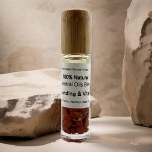 May include: A clear glass essential oil roller bottle with a wooden cap. The bottle contains red jasper stones and a blend of essential oils. The label reads "100% Natural Essential Oils Blend" and lists ingredients.