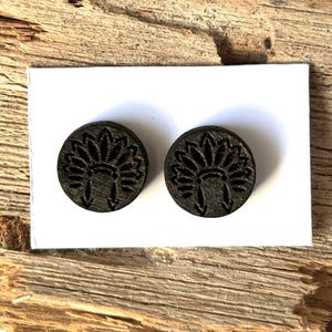 May include: Two dark brown, round earrings with a Native American headdress design. The earrings are made of wood and are displayed on a white card, set against a rustic wooden background.