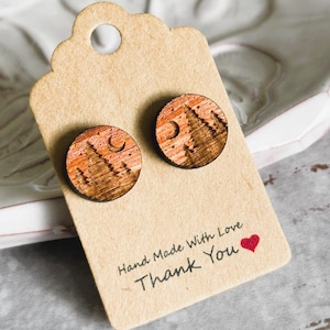 May include: Round wooden stud earrings with a mountain and moon design. The earrings have a natural wood grain finish and a detailed landscape illustration. The earrings are displayed on a tag that reads "Hand Made With Love Thank You" with a small red heart.