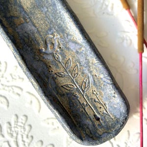 Könnte beinhalten: Ein handgefertigter Keramik-Räucherstäbchenhalter in Blau- und Goldtönen. Der Halter hat ein geprägtes Blumenmuster und ein kleines Loch für Räucherstäbchen. Zwei Räucherstäbchen mit rosa Spitzen sind sichtbar.