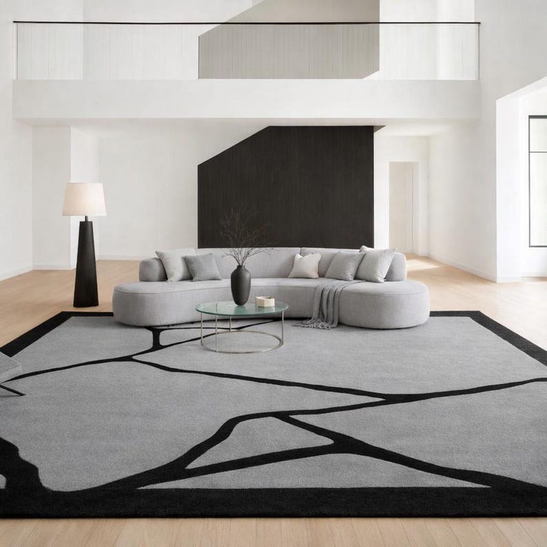 May include: A modern living room featuring a large gray rug with a black geometric design. A gray sectional sofa with throw pillows sits on the rug. A glass-topped coffee table and a black floor lamp complete the space.