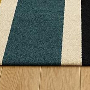 May include: A rectangular rug with a striped pattern. The rug features alternating stripes of rust, yellow, teal, black, and cream. The rug is placed on a light-colored wooden floor.