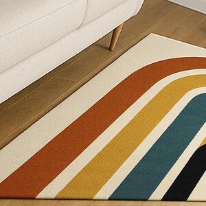 May include: A retro-style area rug with a cream-colored background and bold, curved stripes in shades of orange, yellow, teal, and black. The rug is placed on a hardwood floor, with a portion of a white sofa visible in the upper left corner.