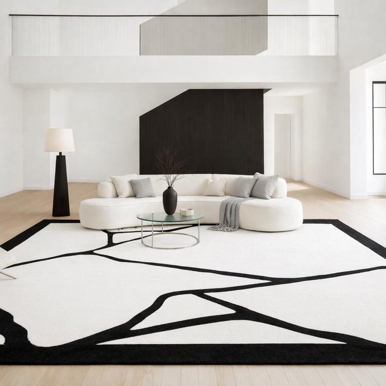 May include: A modern living room featuring a large white sofa with gray pillows, a round glass coffee table, and a black floor lamp. A large white and black rug with an abstract design covers the floor. The walls are white with a black accent wall.