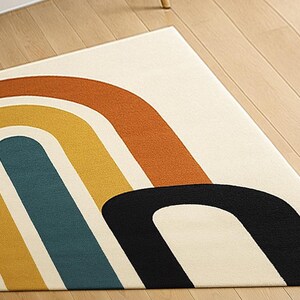 May include: A rectangular rug with a cream-colored background and a retro rainbow design. The rainbow arches are in shades of orange, yellow, teal, and black. The rug is placed on a light-colored wooden floor.