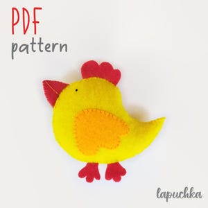 Puede incluir: Una manualidad de fieltro amarillo con forma de pollo, con cresta y pico rojos, ala naranja y patas rojas. El texto PDF pattern aparece en rojo y gris, y el texto lapuchka en gris. El pollo tiene un pequeño ojo negro y costuras visibles.
