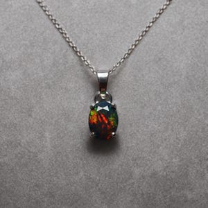 May include: A silver necklace with an oval-shaped pendant. The pendant features a dark opal with flashes of red, green, and yellow. The opal is set in a silver bezel and hangs from a delicate silver chain.