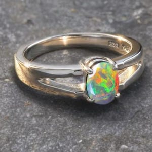 Op de afbeelding: Een zilveren ring met een ovale opaal edelsteen. De opaal toont een levendig kleurenpalet, waaronder groen, oranje en rood. De ring heeft een split-schacht ontwerp.