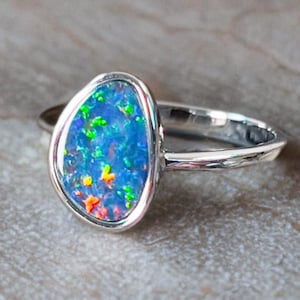 May include: A silver ring featuring a vibrant opal gemstone. The opal has a blue base color with flashes of green, orange, and yellow. The gemstone is set in a bezel setting, and the ring band is smooth and polished.