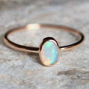 Könnte beinhalten: Roségoldener Ring mit einem ovalen Opal-Edelstein. Der Opal zeigt ein Farbspiel mit schillernden Blau-, Grün- und Gelbtönen. Der Ring hat ein dünnes Band, was zu seinem eleganten Design beiträgt.