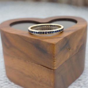 May include: A gold band ring with a continuous row of small, dark blue gemstones. The ring is set on a heart-shaped wooden box with a natural wood grain finish. The ring is a classic eternity style.