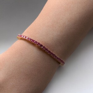 May include: A gold bracelet adorned with numerous small, vibrant red gemstones. The bracelet is set on a wrist, showcasing the jewelry's design and color. The gemstones are closely set, creating a continuous line of color.