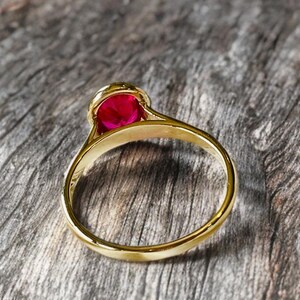 K&ouml;nnte beinhalten: Ein goldener Ring mit einem gro&szlig;en, runden, leuchtend roten Edelstein. Der Ring hat ein glattes, poliertes Band, und der Edelstein ist in einer detaillierten, verzierten Fassung gefasst. Der Ring wird auf einer strukturierten Holzoberfl&auml;che pr&auml;sentiert.