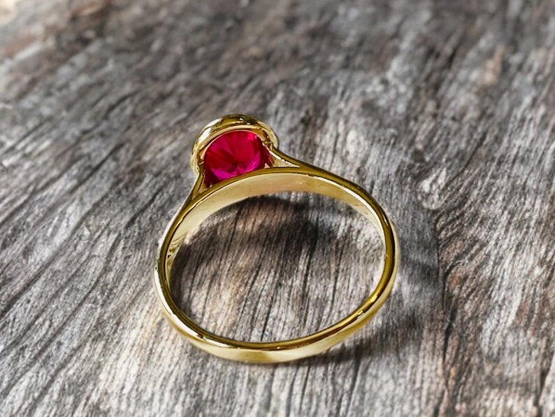 K&ouml;nnte beinhalten: Ein goldener Ring mit einem gro&szlig;en, runden, leuchtend roten Edelstein. Der Ring hat ein glattes, poliertes Band, und der Edelstein ist in einer detaillierten, verzierten Fassung gefasst. Der Ring wird auf einer strukturierten Holzoberfl&auml;che pr&auml;sentiert.