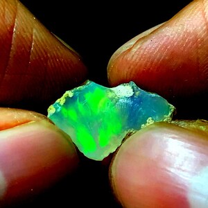 Peut inclure: Une pierre précieuse d'opale verte et bleue de forme brute et naturelle. L'opale est tenue entre deux doigts.