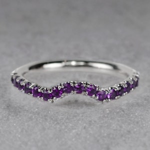 May include: A silver ring with a curved design, set with small, vibrant purple gemstones. The gemstones are evenly spaced along the ring's curve, creating a continuous line of color. The ring is shown against a neutral gray background.