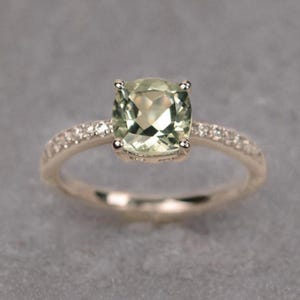 Puede incluir: Un anillo de oro con una gran piedra preciosa verde pálido de talla cuadrada. La banda está adornada con pequeñas piedras de acento transparentes. El anillo se muestra sobre un fondo gris neutro, resaltando los detalles de la joya.