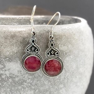 Könnte beinhalten: Ein Paar silberfarbene Hängeohrringe. Jeder Ohrring hat einen runden, tiefroten Edelstein, der in einen dekorativen Rahmen im Filigranstil gefasst ist. Die Ohrringe haben einen Hakenverschluss und sind vor neutralem Hintergrund abgebildet.