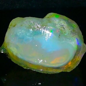 Könnte beinhalten: Ein rauer, durchscheinender, weißer Opal-Edelstein mit irisierenden grünen, blauen und orangefarbenen Blitzen. Der Opal ist unregelmäßig geformt und hat eine glatte, polierte Oberfläche.