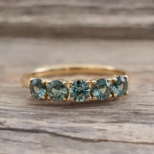 Könnte beinhalten: Ein goldener Ring mit fünf runden, teal-farbenen Edelsteinen. Die Edelsteine sind eng nebeneinander angeordnet. Der Ring wird auf einer Holzoberfläche präsentiert. Das Design ist schlicht und elegant.