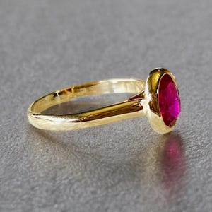 K&ouml;nnte beinhalten: Ein goldener Ring mit einem leuchtenden, oval geformten Rubin-Edelstein. Band und Fassung des Rings sind goldfarben poliert und bilden einen auff&auml;lligen Kontrast zum tiefen Rot des Edelsteins. Der Ring ist ein Schmuckst&uuml;ck.