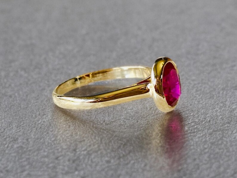 K&ouml;nnte beinhalten: Ein goldener Ring mit einem leuchtenden, oval geformten Rubin-Edelstein. Band und Fassung des Rings sind goldfarben poliert und bilden einen auff&auml;lligen Kontrast zum tiefen Rot des Edelsteins. Der Ring ist ein Schmuckst&uuml;ck.