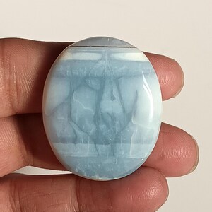 Peut inclure: Pierre précieuse ovale polie. La pierre présente un mélange de teintes bleu clair et blanc, avec de subtiles motifs veinés. Le bijou est tenu dans une main, mettant en valeur sa taille et sa forme. L'aspect général est lisse et brillant.