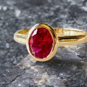 K&ouml;nnte beinhalten: Ein goldener Ring mit einem gro&szlig;en, oval geschliffenen Rubin. Der Rubin hat eine leuchtend rote Farbe und ist in einem glatten, polierten Goldband gefasst. Der Ring wird auf einer strukturierten, grauen Oberfl&auml;che pr&auml;sentiert.