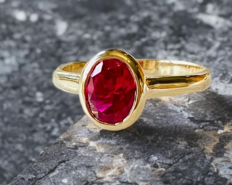 K&ouml;nnte beinhalten: Ein goldener Ring mit einem gro&szlig;en, oval geschliffenen Rubin. Der Rubin hat eine leuchtend rote Farbe und ist in einem glatten, polierten Goldband gefasst. Der Ring wird auf einer strukturierten, grauen Oberfl&auml;che pr&auml;sentiert.