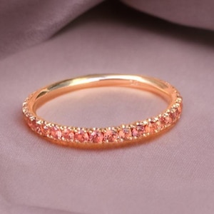 Op de afbeelding: Een goudkleurige ring met een band van kleine, perzik-kleurige edelstenen. De ring staat tegen een zachte, gedempte achtergrond, die het delicate ontwerp en de warme tinten van de ring benadrukt. De edelstenen zijn gelijkmatig verdeeld.