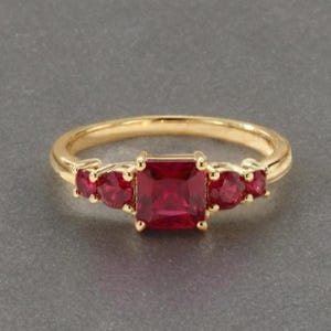 May include: A gold ring featuring a central square-cut red gemstone flanked by two smaller round-cut red gemstones on each side. The band is polished yellow gold, and the setting holds the five red stones in a symmetrical, prong-set arrangement.