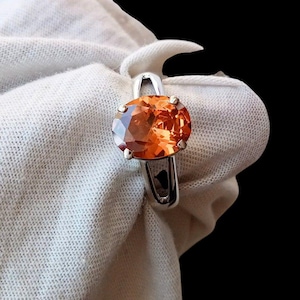 May include: A silver ring featuring a large, oval-cut orange gemstone. The gemstone is the focal point, set in a simple, elegant band. The ring is displayed against a white fabric background.