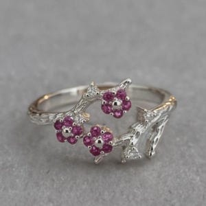 May include: A silver ring featuring a textured branch-like band adorned with three small flowers, each composed of five vibrant pink gemstones. Small, clear, sparkling stones are set at intervals along the silver branches, adding detail.