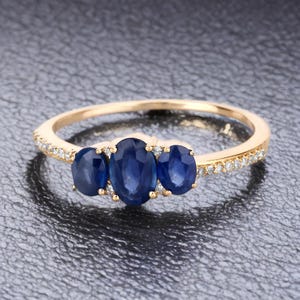 May include: A gold ring featuring three oval, deep blue gemstones. The band is also adorned with small, clear gemstones. The ring is set against a dark, textured background, highlighting the sparkle of the gems.