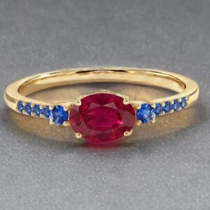 May include: A gold ring featuring a central oval-cut red gemstone flanked by a row of small round blue gemstones on each side of the band. The metal band is polished yellow gold, and the stones are set in a prong setting along the top of the ring.