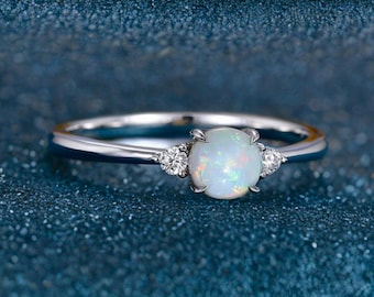 Pode incluir: Um anel de prata com uma pedra de opala redonda e central, ladeada por duas pedras preciosas claras menores. A opala é branca com flashes de cor. O anel está sobre um fundo azul escuro. A opala tem cerca de 0,8 cm de diâmetro.