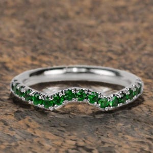May include: A curved silver ring set with a row of vibrant green gemstones. The gemstones are evenly spaced along the top of the ring, creating a continuous line of color. The ring is set against a textured brown background.