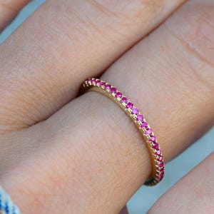 Puede incluir: Un anillo de banda dorado engastado con pequeñas y vibrantes piedras preciosas rosas. El anillo se muestra en un dedo, con las piedras preciosas que rodean toda la banda. El diseño es simple y elegante, con un enfoque en el color brillante de las piedras.