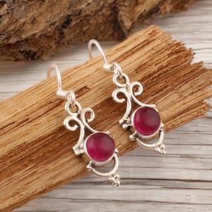 Könnte beinhalten: Ein Paar silberne Ohrhänger mit dekorativem Design. Jeder Ohrring hat einen runden, tiefrosa Edelstein, der in einen silbernen Rahmen gefasst ist. Die Ohrringe werden auf einem Holzstück präsentiert.