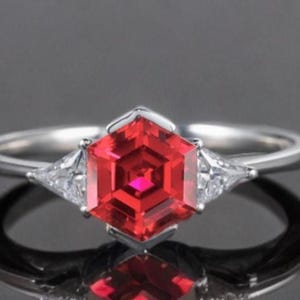 May include: A silver ring with a striking, hexagonal, red gemstone at its centre. Two smaller, triangular, clear gemstones are either side of the main stone. The ring is displayed on a dark, reflective surface.