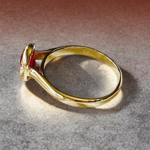K&ouml;nnte beinhalten: Ein goldener Ring mit einem roten Edelstein. Der Ring hat ein glattes, poliertes Band und eine L&uuml;nettenfassung f&uuml;r den Edelstein. Der Ring ist mit "14k" markiert.