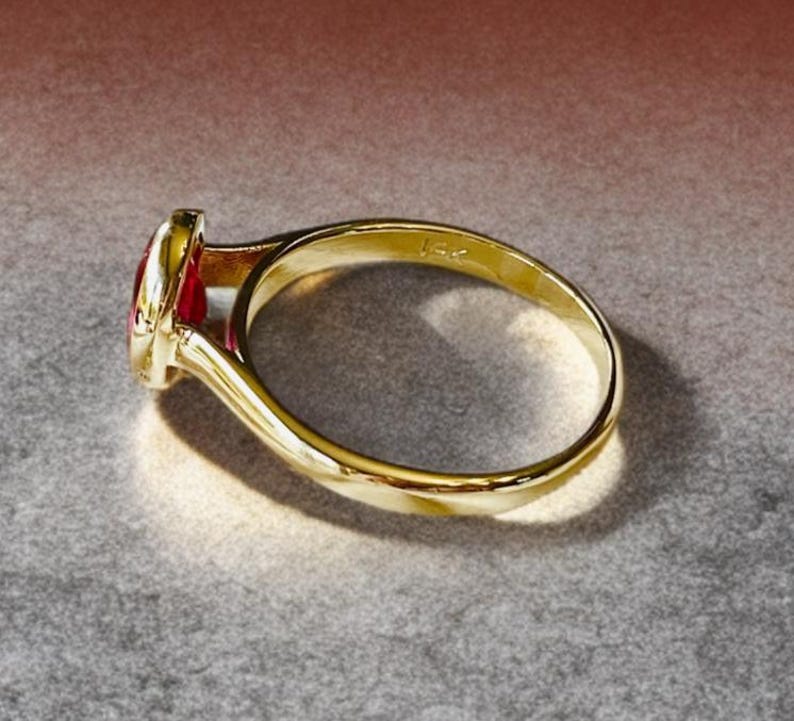 K&ouml;nnte beinhalten: Ein goldener Ring mit einem roten Edelstein. Der Ring hat ein glattes, poliertes Band und eine L&uuml;nettenfassung f&uuml;r den Edelstein. Der Ring ist mit "14k" markiert.