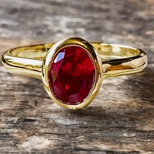 K&ouml;nnte beinhalten: Ein goldener Ring mit einem gro&szlig;en, oval geschliffenen Rubin-Edelstein. Der Rubin hat eine tiefrote Farbe und ist in eine goldene L&uuml;nette gefasst. Der Ring liegt auf einer Holzoberfl&auml;che.
