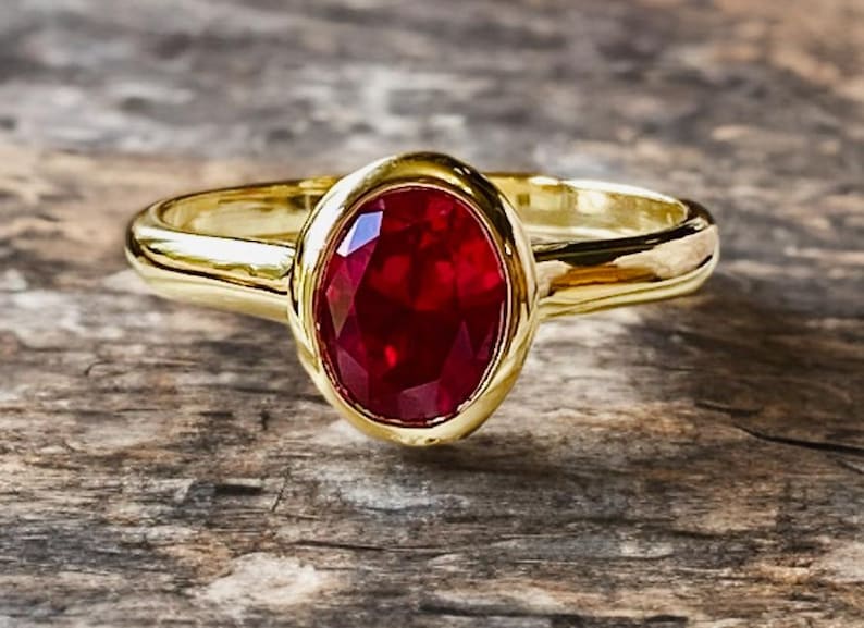 K&ouml;nnte beinhalten: Ein goldener Ring mit einem gro&szlig;en, oval geschliffenen Rubin-Edelstein. Der Rubin hat eine tiefrote Farbe und ist in eine goldene L&uuml;nette gefasst. Der Ring liegt auf einer Holzoberfl&auml;che.